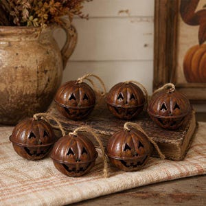 May include: A set of six round, rusted metal jingle bells shaped like jack-o'-lanterns. Each bell features cut-out triangular eyes and a jagged mouth, with a natural jute twine loop attached to the top for hanging, showcasing a weathered finish.
