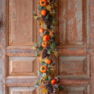 May include: A decorative garland featuring bright orange persimmon fruits, dark brown pinecones, and green leaves. The arrangement includes small clusters of tiny orange berries interspersed throughout the foliage, creating a natural, textured appearance.