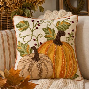 May include: A square throw pillow with an off-white fabric base featuring an embroidered design of two pumpkins. One pumpkin is large and golden-yellow, while the smaller one is beige. Green leaves, brown stems, and small red berries accent the design.