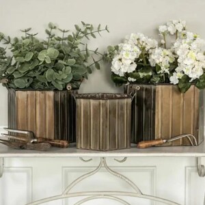 May include: Three decorative metal planters with vertical ribbed detailing, filled with greenery and white flowers. The planters are in varying sizes and sit on a white shelf. Gardening tools are placed in front of the planters.
