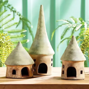 May include: Three ceramic fairy houses with green roofs and brown walls. The houses have small windows and doors. The houses are arranged on a wooden surface.