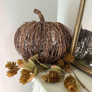 May include: A decorative pumpkin crafted from brown intertwined twigs and vines, featuring a rounded shape with a prominent stem. A sprig of dried green and brown leaves and small pinecone-like clusters rests at the base of the pumpkin.