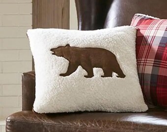 May include: A white faux fur throw pillow with a brown bear silhouette appliqué. The pillow is sitting on a brown leather chair.