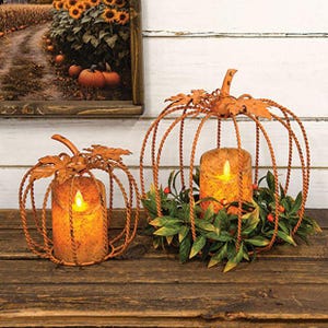 May include: Two orange metal pumpkin-shaped candle holders with twisted wire frames and decorative leaves at the top. Each frame contains a cylindrical LED candle with a warm yellow glow. The larger pumpkin is surrounded by green leaves and red berries.