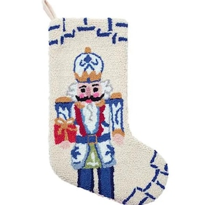 May include: A Christmas stocking with a cream-colored background and a detailed design of a nutcracker soldier. The nutcracker is dressed in blue, white, red, and gold, holding a small red gift. The stocking has a loop for hanging.