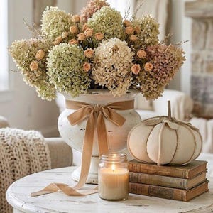 May include: A white ceramic urn filled with dried green and beige hydrangea flowers and small peach-colored roses. A tan velvet ribbon is tied around the urn. Beside it sits a fabric pumpkin, a stack of three vintage books, and a lit glass candle.