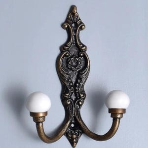 May include: A double hook wall hanger with an ornate, black metal design and two white ceramic knobs.