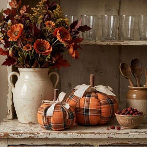May include: A cream-colored ceramic vase filled with orange and dark red artificial flowers, two fabric pumpkins with an orange plaid pattern and beige polka-dot ribbon bows, and a small bowl containing red berries on a distressed white wooden surface.