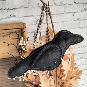 May include: A black burlap crow decoration with white stitching, perched on a fall-themed wreath. The wreath includes dried leaves, twigs, and decorative accents. The crow is a rustic, handcrafted item, perfect for autumn or Halloween decor.