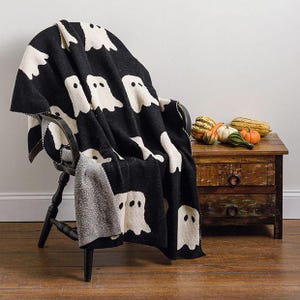 May include: A black knit throw blanket featuring a repeating pattern of white ghost figures. The blanket is draped over a dark wooden chair. The fabric has a visible woven texture with a contrasting white reverse side visible at the folded edge.