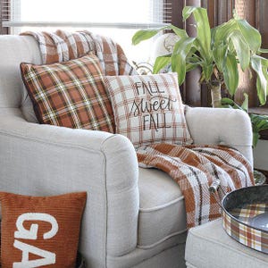 May include: A light gray armchair featuring a brown and white plaid throw blanket, a brown plaid throw pillow, and a white throw pillow with a brown plaid grid pattern and the black text "FALL sweet FALL" printed on the front surface.