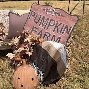 May include: A decorative metal sign with a distressed red finish and black text reading PUMPKIN FARM, featuring a floral motif. It is displayed with a plaid blanket, dried oak leaves, and a light orange ceramic pumpkin with star cutouts.