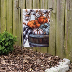 May include: A rectangular garden flag hanging on a black metal stand, featuring a printed illustration of orange, blue, and white pumpkins in a dark wooden barrel, accented with cotton bolls, wheat stalks, and a black and white buffalo check cloth.