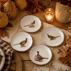 May include: A set of four round white ceramic plates with scalloped edges, each featuring a detailed illustration of a pheasant bird in natural colors. The plates have a thin brown rim and are arranged on a textured fabric surface.