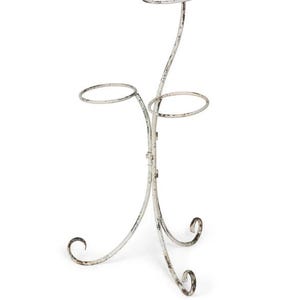 May include: A distressed white metal plant stand with three circular holders. The stand has a curved design with decorative scrollwork at the base. The stand is designed to hold multiple plants.