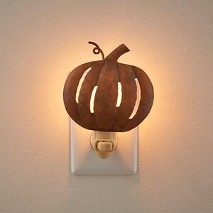 May include: A plug-in night light featuring a rustic, brown metal pumpkin silhouette with vertical cutouts that allow warm light to shine through. The pumpkin has a small curled stem at the top and is mounted on a standard white plastic base.