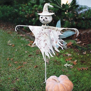 May include: A metal scarecrow garden stake featuring a white, distressed finish with rust-colored speckles. The figure wears a pointed hat and has a carved pumpkin-style head with triangular eyes and a jagged mouth, standing above a large pumpkin.