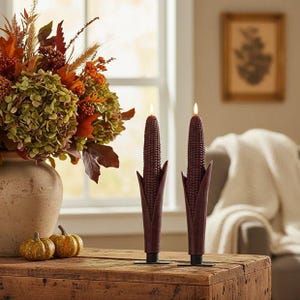 May include: Two dark burgundy taper candles shaped like ears of corn, each featuring a textured kernel surface and a husk base. The candles are mounted on small black rectangular stands and topped with a small, glowing artificial flame light.