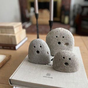 May include: Three gray, rounded, porous stones arranged on a book. Each stone features two small circular indentations for eyes and one larger circular indentation for a mouth, creating a simple face. The stones have a rough, natural texture.