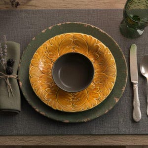 May include: A place setting featuring a large, dark green charger plate with a distressed edge, topped with a yellow, leaf-patterned dinner plate and a small, solid dark brown bowl. A green napkin, a silver knife, a silver spoon, and a green glass.