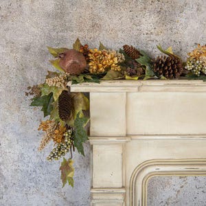 May include: An autumn-themed garland draped over a cream-colored mantelpiece, featuring artificial green and brown maple leaves, brown pinecones, clusters of small green berries, dried yellow flowers, and a single textured brown pomegranate.