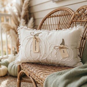May include: A rectangular off-white throw pillow with frayed edges featuring two embroidered pumpkin outlines. Each pumpkin has a small tan tag tied with twine; the left tag reads sweater weather and the right tag reads cozy.