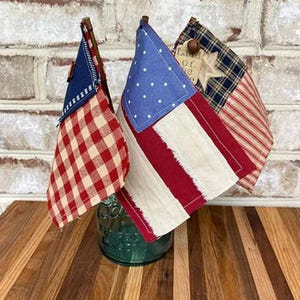 May include: Three decorative American flags with patriotic designs. One flag features a blue field with white dots, red and white stripes. Another has a red and white checkered pattern, and the third has a star and stripe design. They are displayed in a green jar.