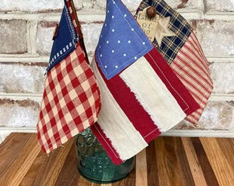May include: Three decorative American flags with patriotic designs. One flag features a blue field with white dots, red and white stripes. Another has a red and white checkered pattern, and the third has a star and stripe design. They are displayed in a green jar.