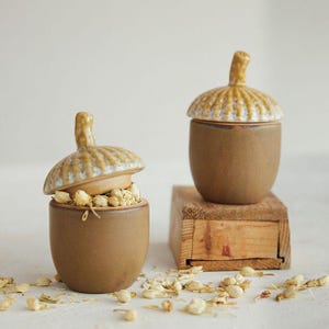 May include: Two ceramic jars shaped like acorns with smooth brown bases and textured, glossy tan lids. One jar is open, revealing dried jasmine flowers inside, while the other is closed. Scattered dried jasmine flowers surround the jars.