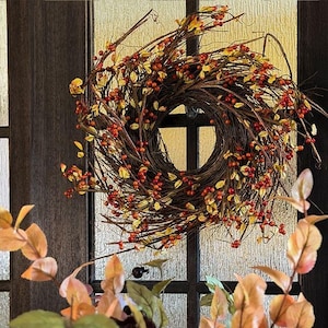 May include: A circular wreath constructed from dark brown intertwined twigs, adorned with clusters of small red berries and scattered yellow and orange leaves. The wreath is mounted on a dark frame with glass panels, featuring a natural aesthetic.