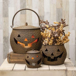May include: Three brown metal jack-o'-lantern containers with hammered textures and cutout faces. The largest has a handle and lid, the smallest is a lidded pot, and the third is an open-top bucket filled with artificial yellow berries and leaves.