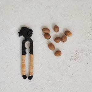 May include: A black cast iron nutcracker with a woven rattan handle, and a group of brown nutmeg seeds on a white background.