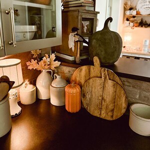 May include: A collection of kitchen decor featuring two light-colored ceramic jugs, a small orange pumpkin-shaped jar, two wooden pumpkin-shaped cutting boards, and a large dark gray decorative pumpkin, all arranged on a dark countertop.