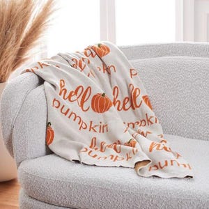 May include: A cream-colored throw blanket draped over a gray armchair. The blanket features the words "hello pumpkin" in orange script, along with orange pumpkin illustrations. The blanket is a cozy fall-themed home decor item.