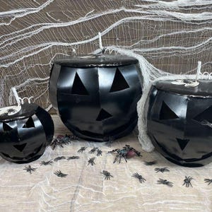 May include: Three black metal jack-o'-lanterns with triangular eyes and noses, featuring cut-out mouths. The pumpkins are decorated with small plastic spiders and white faux spiderwebs draped over and around the containers.
