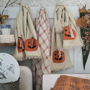 May include: Two cream-colored fabric towels featuring orange pumpkin appliqués with black stitched faces and frayed edges, hanging alongside a white and orange plaid fabric cloth. The towels are displayed on a white wooden rack.