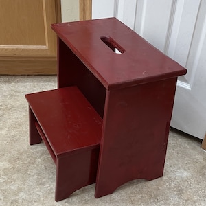 May include: A two-step wooden stool painted in a deep red color. The top step has a cutout for easy carrying.