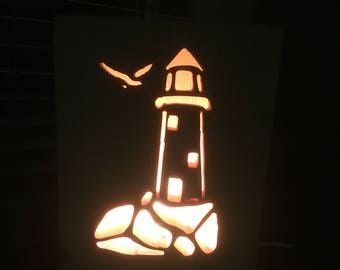May include: A wooden cutout of a lighthouse with a bird flying overhead, illuminated from behind. The lighthouse is on a rocky base.