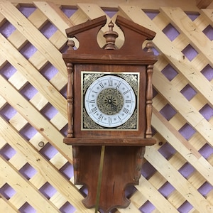 May include: A wooden wall clock with a pendulum. The clock face has Roman numerals and a gold trim. The clock is hanging on a wooden lattice wall.