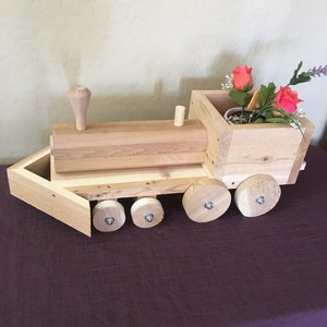 May include: A wooden toy train with a planter box on the back. The train is painted in a natural wood color and has four wheels. The planter box is filled with a small potted plant and pink flowers.