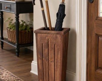 May include: A tall, rectangular wooden umbrella stand with a dark brown finish and vertical grooved detailing on the front panel. The stand holds two wooden walking canes with curved handles and one black umbrella with a curved handle.