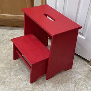 May include: A red wooden step stool with a cutout in the top step. The stool has two steps.