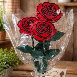 May include: A bouquet of three red rose cutouts with black outlines, wrapped in clear cellophane. The stems are green, and the bouquet is tied with twine and a small tag. The roses are a vibrant red color.