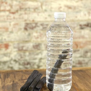 May include: A clear plastic water bottle with a white cap, filled with water and a black charcoal stick inside. A pile of black charcoal sticks are on a wooden surface.