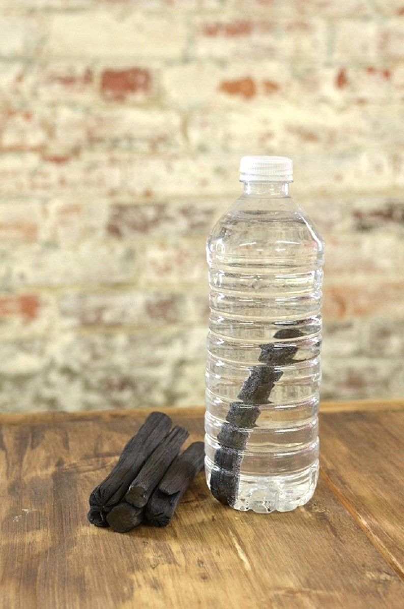 May include: A clear plastic water bottle with a white cap, filled with water and a black charcoal stick inside. A pile of black charcoal sticks are on a wooden surface.