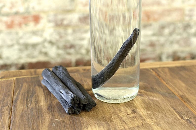 May include: A glass jar filled with water and a long, thin black charcoal stick. A bundle of four smaller charcoal sticks are on a wooden surface.