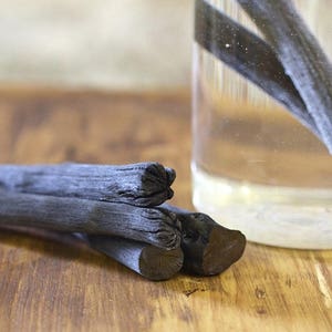 May include: Three black charcoal sticks on a wooden surface. The sticks are stacked on top of each other. A glass jar with water and more charcoal sticks is in the background.