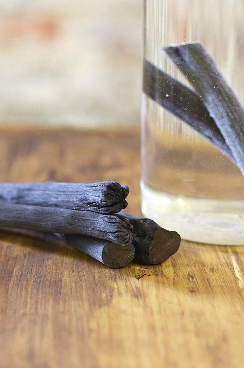May include: Three black charcoal sticks on a wooden surface. The sticks are stacked on top of each other. A glass jar with water and more charcoal sticks is in the background.