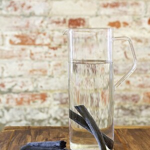May include: A clear glass pitcher filled with water and containing several black charcoal sticks. The pitcher is sitting on a wooden table.