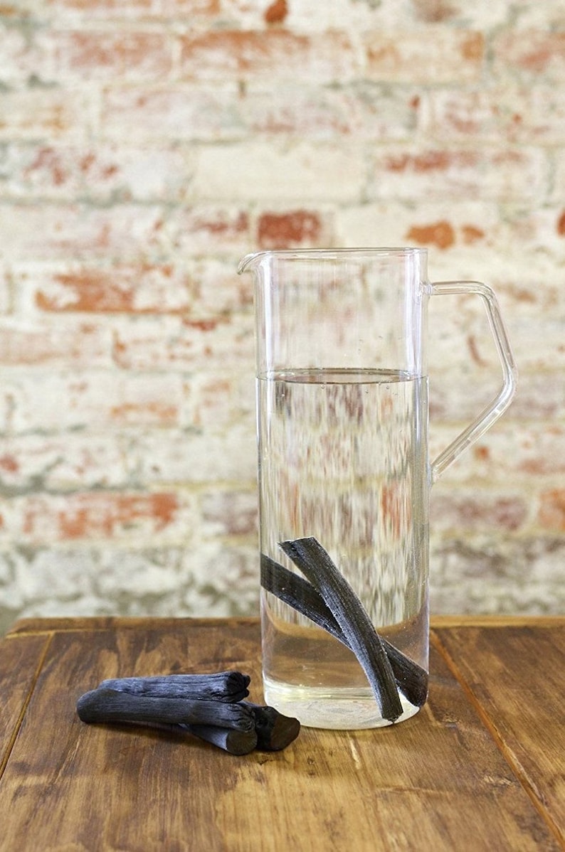 May include: A clear glass pitcher filled with water and containing several black charcoal sticks. The pitcher is sitting on a wooden table.